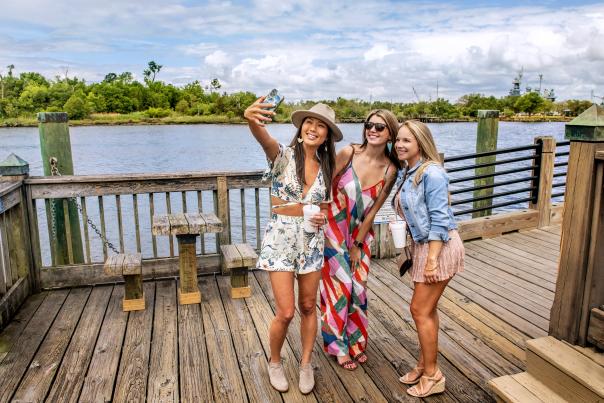 A group of girlfriends taking a selfie on Wilmington Riverwalk with the Battleship NORTH CAROLINA behind them